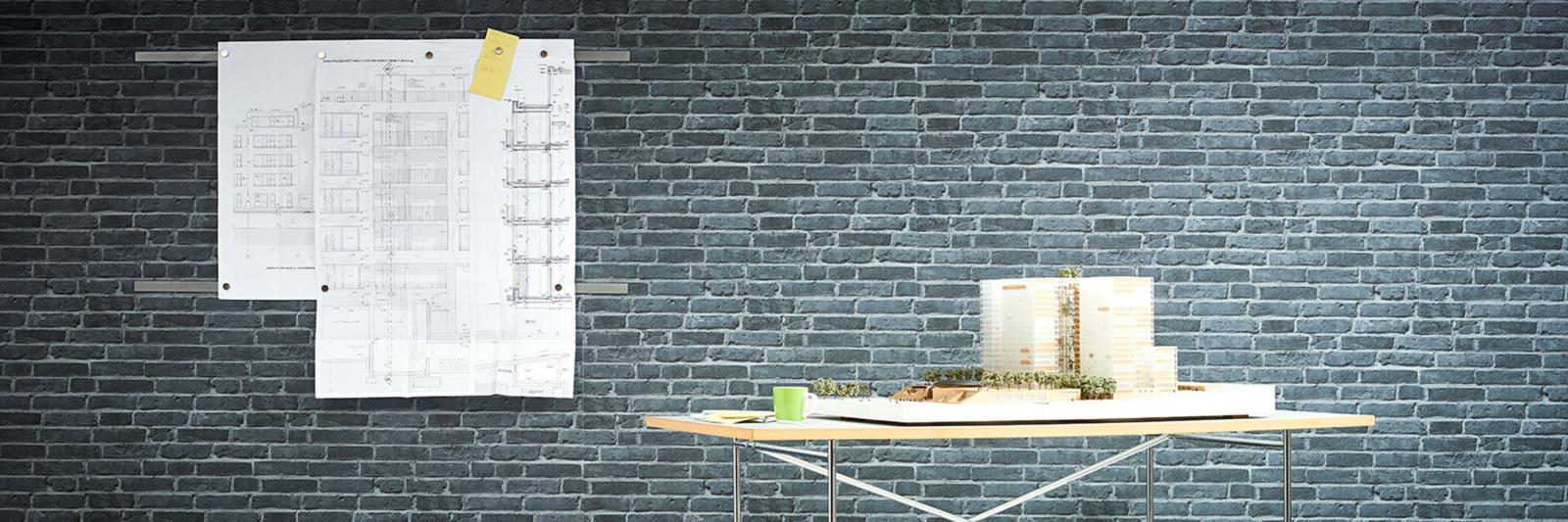 CAD + Reprofolien. Modell eines Gebäudes auf einem Schreibtisch und eine Konstruktionszeichnung auf einer grauen Wand.